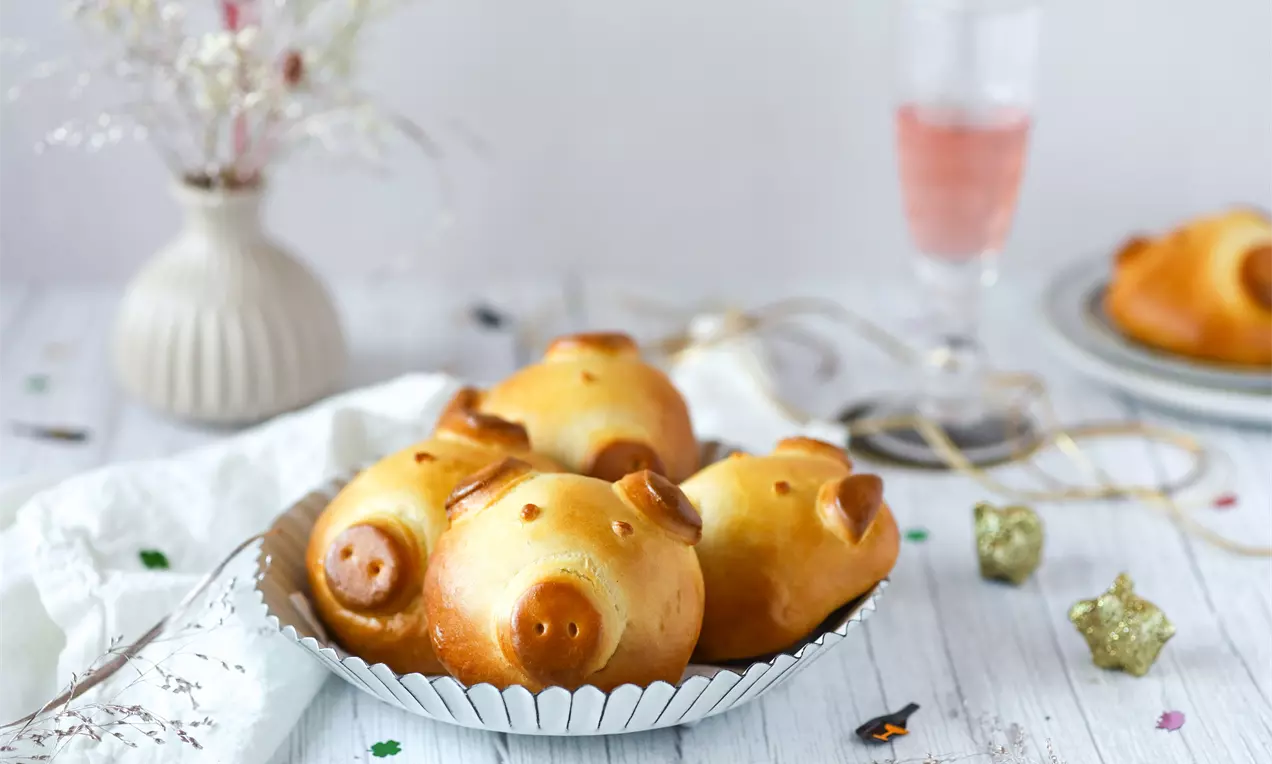 Goldbraune Glücksschweinchen aus Hefeteig mit glänzender Oberfläche in einer silbernen Schale, arrangiert auf einem hellen Holztisch mit Dekoration