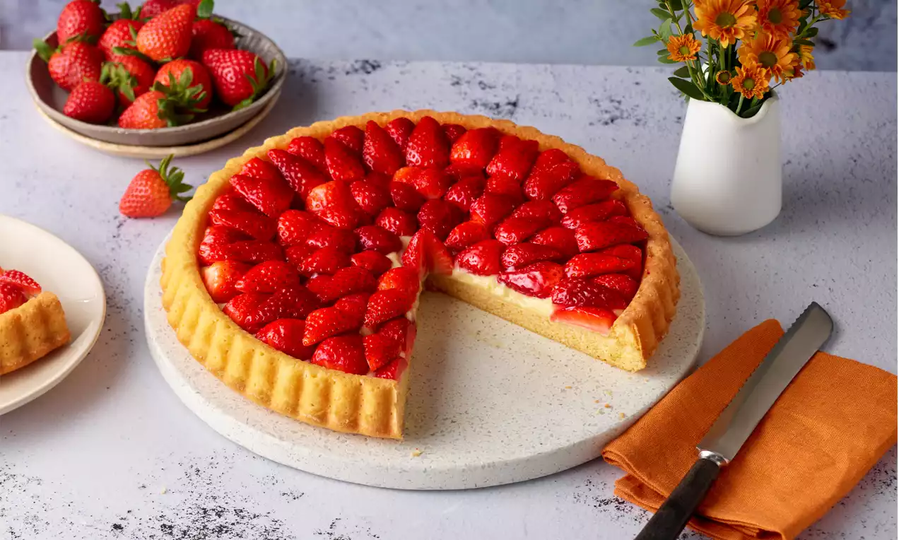 Erdbeerkuchen mit glänzend roten, dekorativ angeordneten Erdbeerscheiben auf hellem Boden, serviert auf rundem Teller mit Messer und Stoffserviette