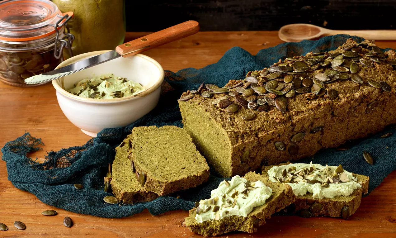 Goldbraunes Kürbisbrot mit knuspriger Kruste und Kürbiskernen bestreut, aufgeschnitten auf dunklem Tuch mit Brotaufstrich und Holzlöffel