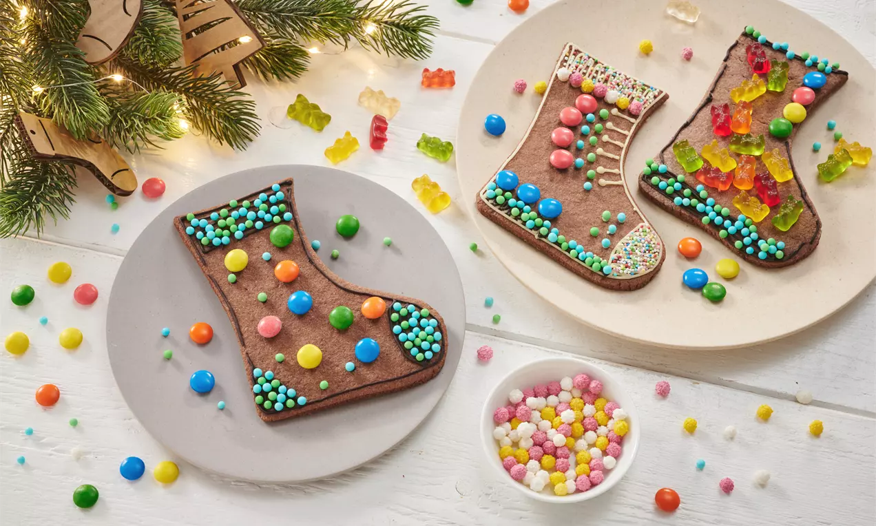 Braune Nikolausstiefel aus Teig, verziert mit bunten Zuckerperlen und Gummibärchen, auf weißen und grauen Tellern, umgeben von Dekoration