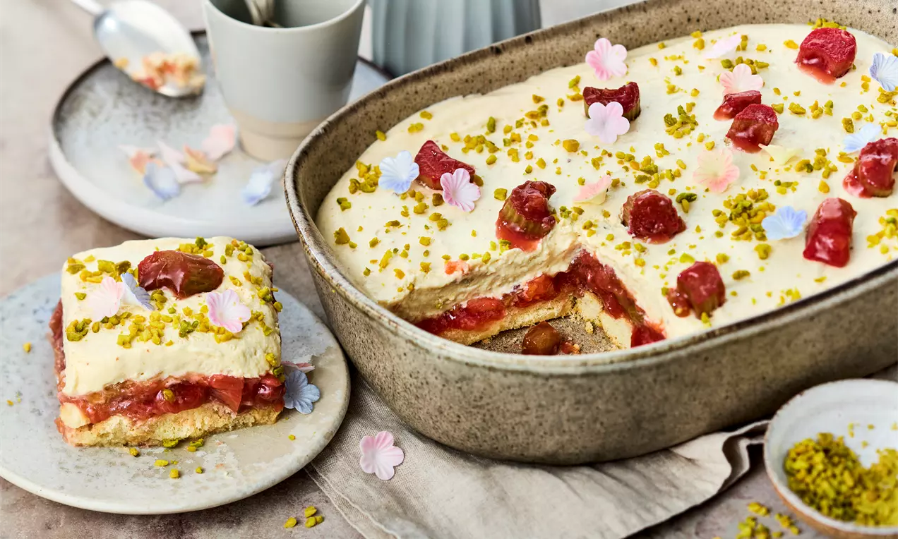 Rhabarber-Tiramisu mit heller Cremeschicht, roten Rhabarberstücken, grünen Pistazien und Blüten dekoriert in grauer Form und auf einem Teller