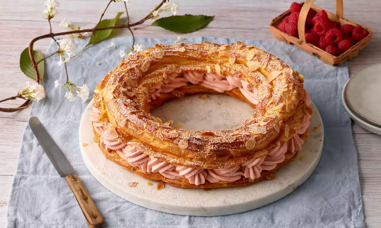 Goldbrauner Brandteigkranz mit rosa Himbeercremefüllung, bestreut mit gehobelten Mandeln auf heller runder Platte, daneben frische Himbeeren
