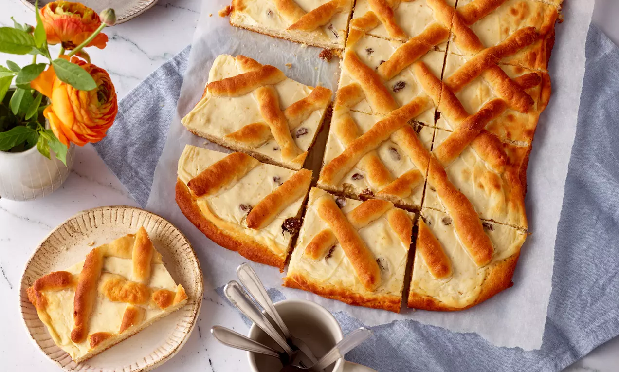 Quadratische Topfenschnitten mit goldbrauner Teiggitterdecke, hellem Belag und Rosinen auf Backpapier, serviert mit Besteck auf hellem Geschirr