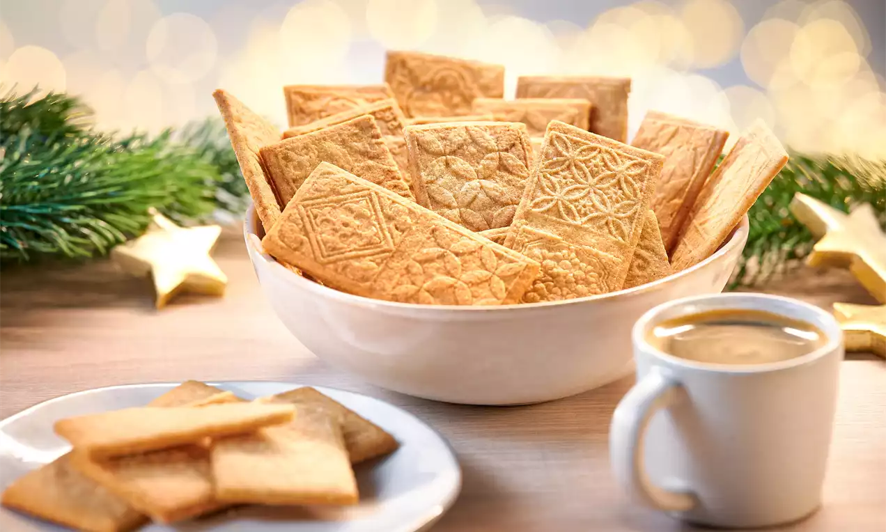 Butter-Spekulatius mit goldbrauner Oberfläche und filigranen Mustern in einer weißen Schale, daneben Tasse Kaffee und Teller mit Keksen