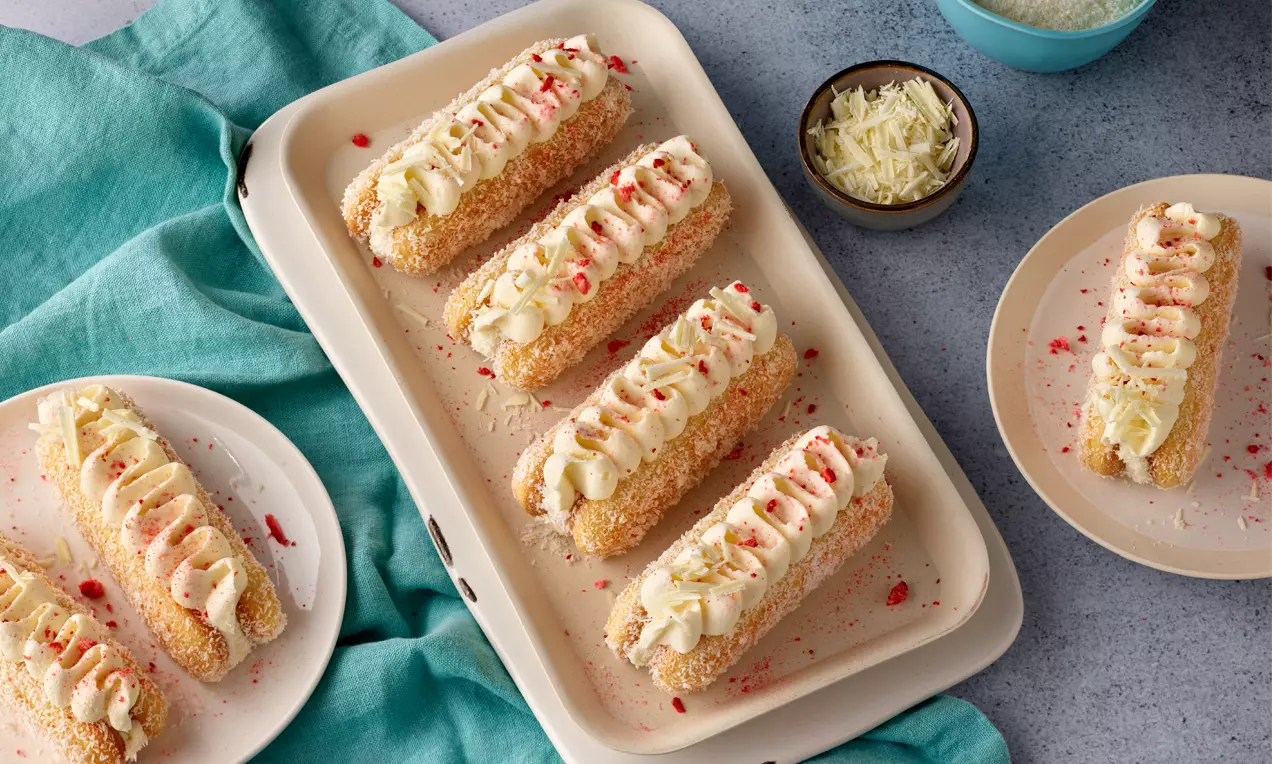 Längliche Erdbeer-Tiramisu-Törtchen mit goldbrauner Keks-Hülle, cremigen Sahnetupfen und roten Erdbeer-Streuseln auf weißem Teller