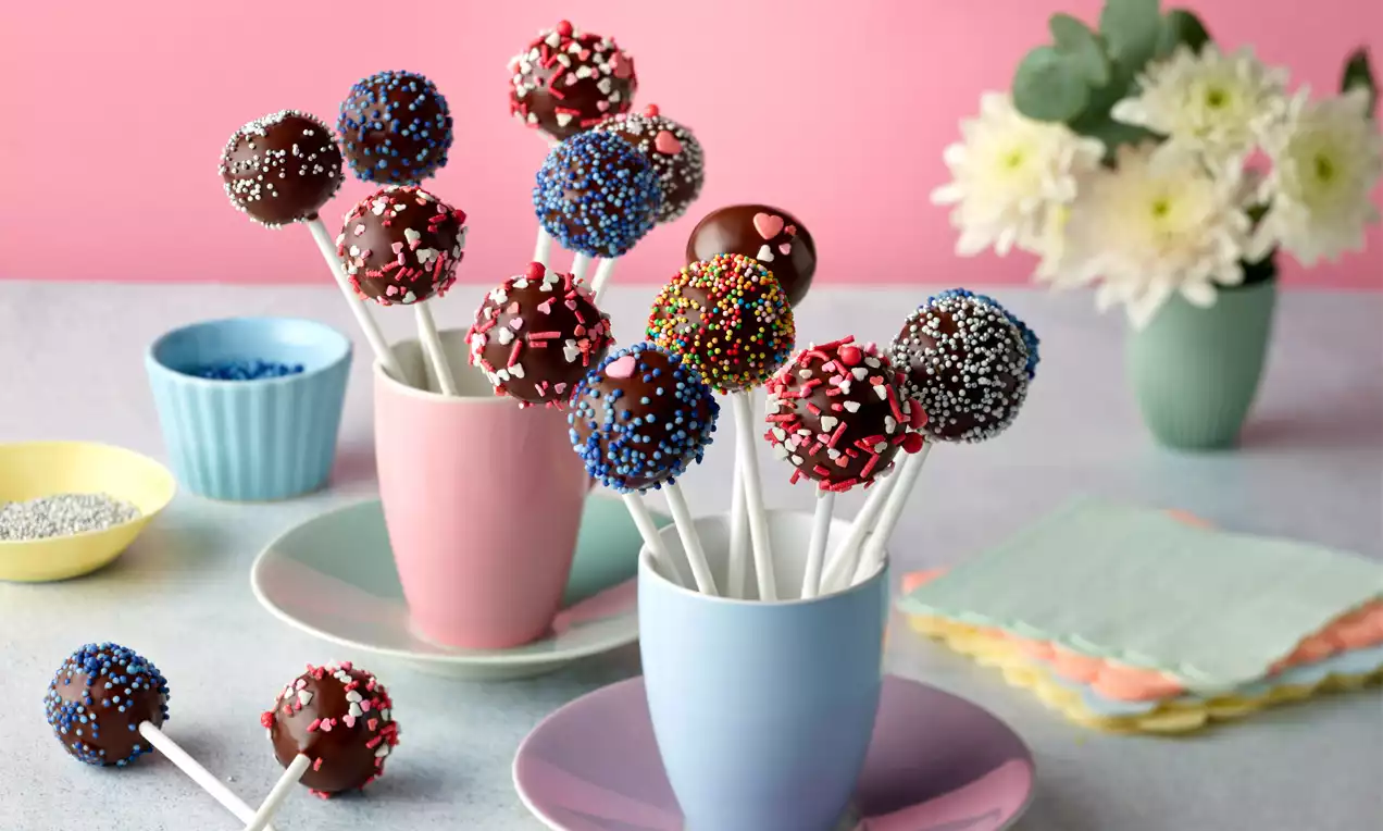 Schoko-Cake-Pops mit glänzender Schokolade, bunt bestreut auf weißen Stielen in farbigen Tassen, umgeben von pastellfarbenem Geschirr und Blumen