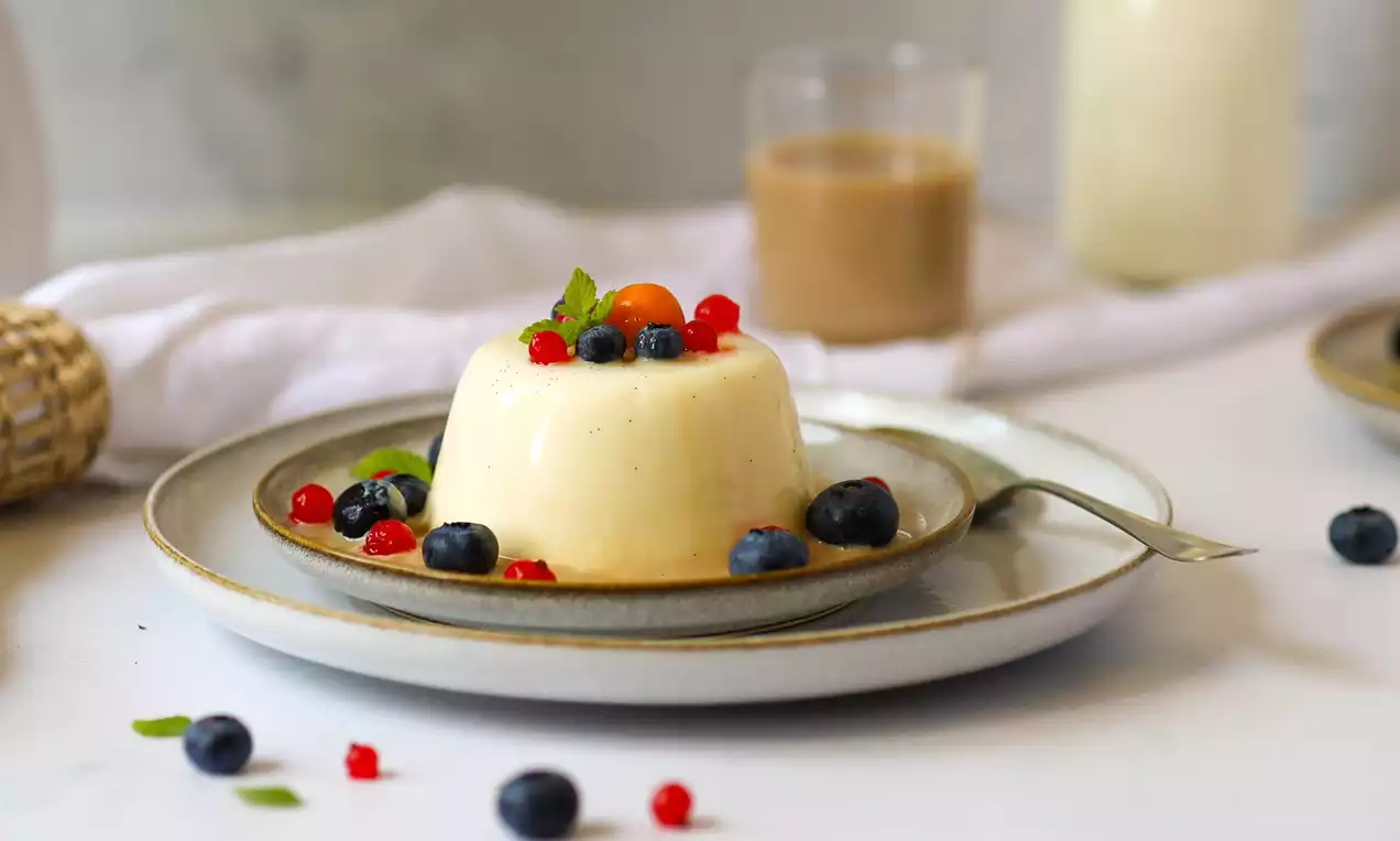 Vanillefarbene Panna Cotta mit glatter Oberfläche, garniert mit roten und blauen Beeren sowie Minzblättern, serviert auf grauem Teller