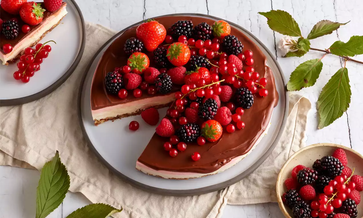 Runde Torte mit glänzendem Überzug aus Schokolade, darauf frische rote und schwarze Beeren, serviert auf einem weißen Teller mit Leinentuch