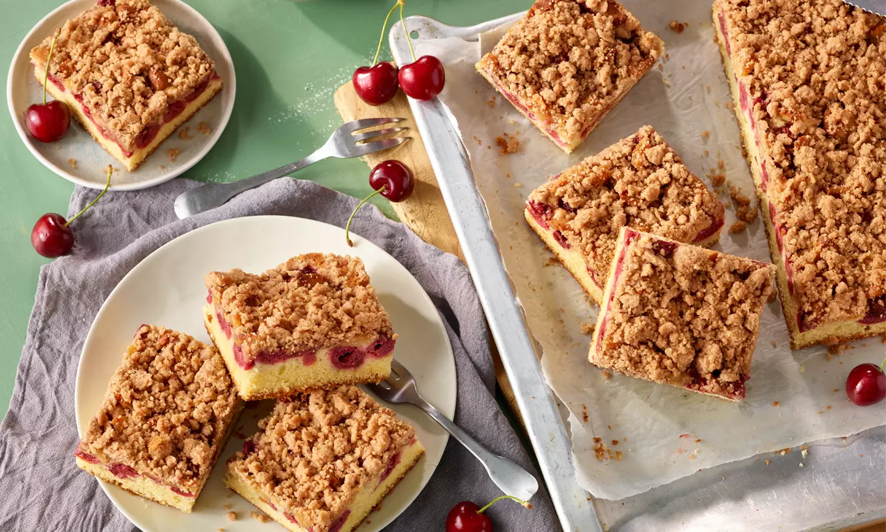 Goldbraune Streusel auf saftigem gelbem Teig mit roten Kirschen, angerichtet auf einem weißen Teller und einem Blech mit frischen Kirschen