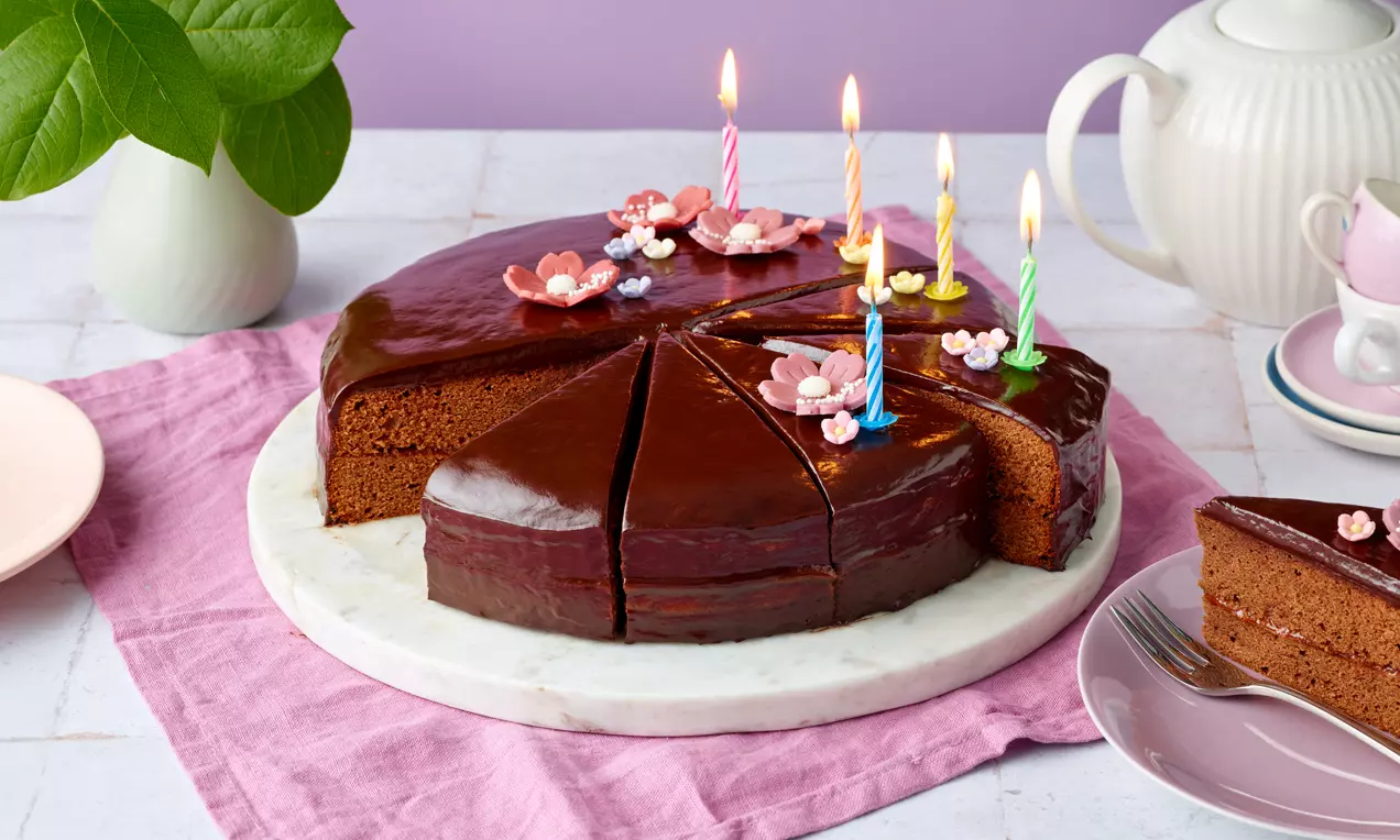 Sacher-Torte mit glänzender dunkler Schokoladenglasur, Kerzen und dekorativen Blüten auf Marmorplatte mit rosa Stoff drapiert