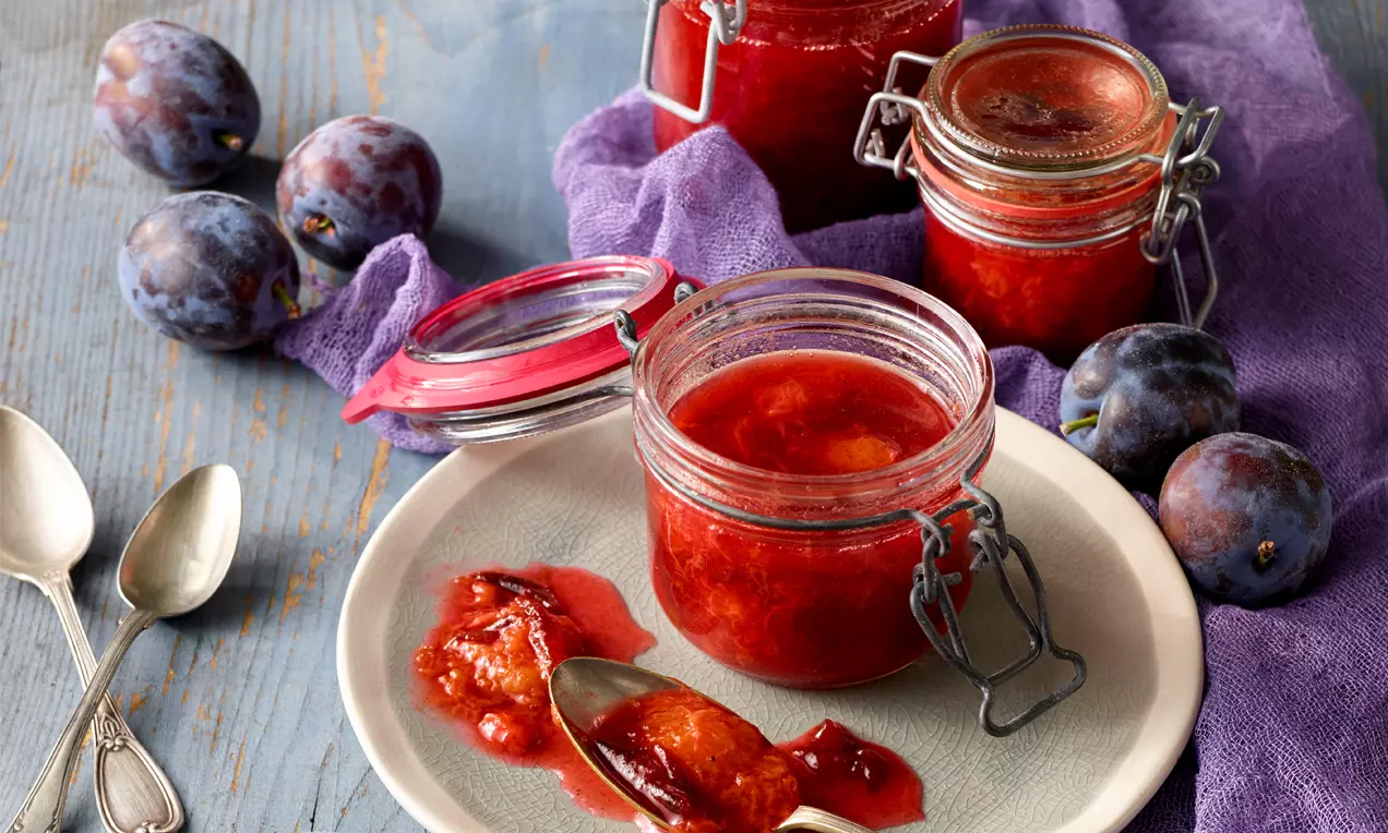 Zwetschkenröster in einem offenen Glas mit leuchtend roter Farbe, daneben Löffel mit Fruchtstücken auf einem Teller, umgeben von frischen Zwetschken