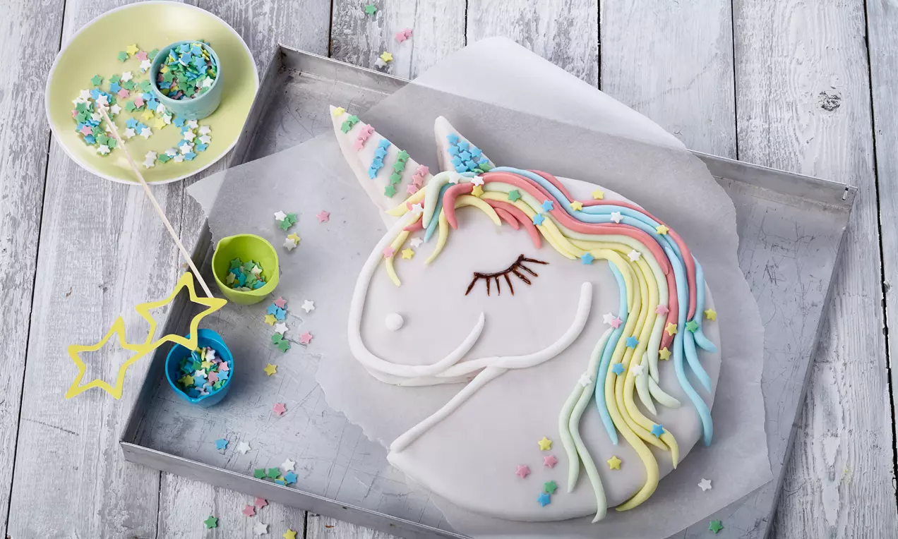 Einhornkuchen in weißer Glasur mit bunter Zuckerdekoration, geschwungener Mähne und detailreich verziertem Horn auf Blechpapier