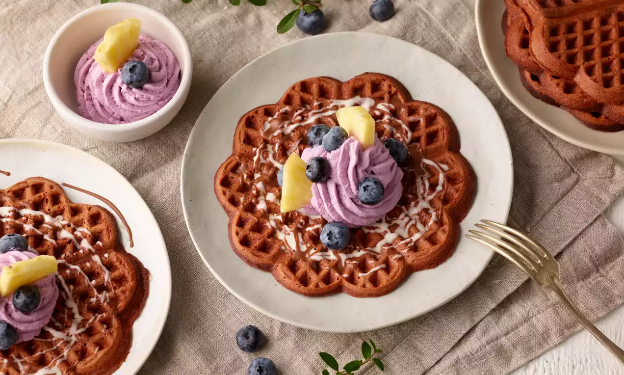 Schokoladenbraune Waffeln mit zarter Heidelbeercreme, frischen Blaubeeren und Ananasstücken auf weißem Teller, mit goldener Gabel dekoriert