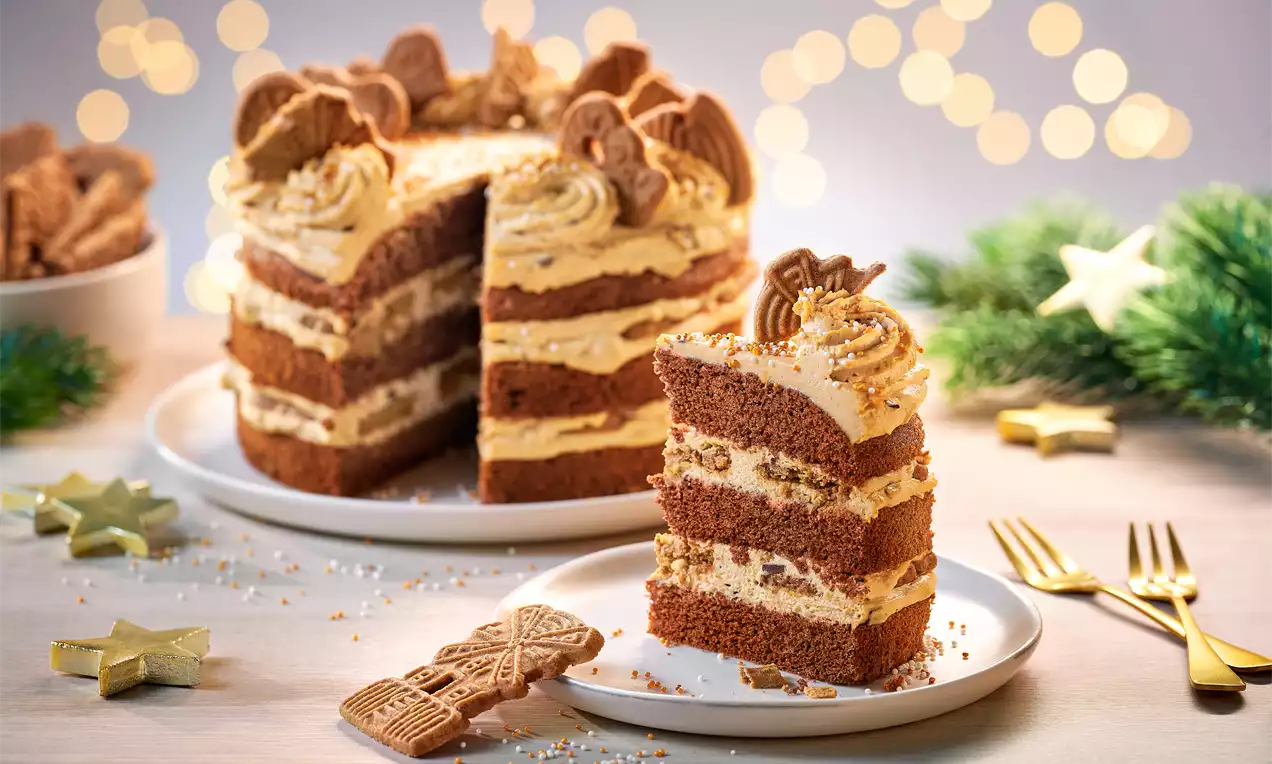 Spekulatius-Törtchen mit braunen Biskuitschichten, heller Creme, Spekulatius-Deko und Streuseln auf weißem Teller vor festlicher Kulisse