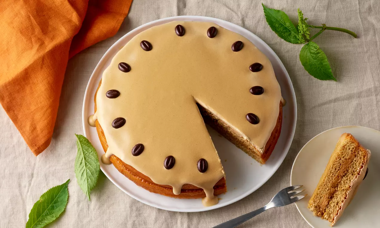 Runde Kaffeetorte mit beigem Zuckerguss, dekoriert mit dunklen Schokobohnen auf weißem Teller, daneben ein Stück auf Untertasse mit Gabel