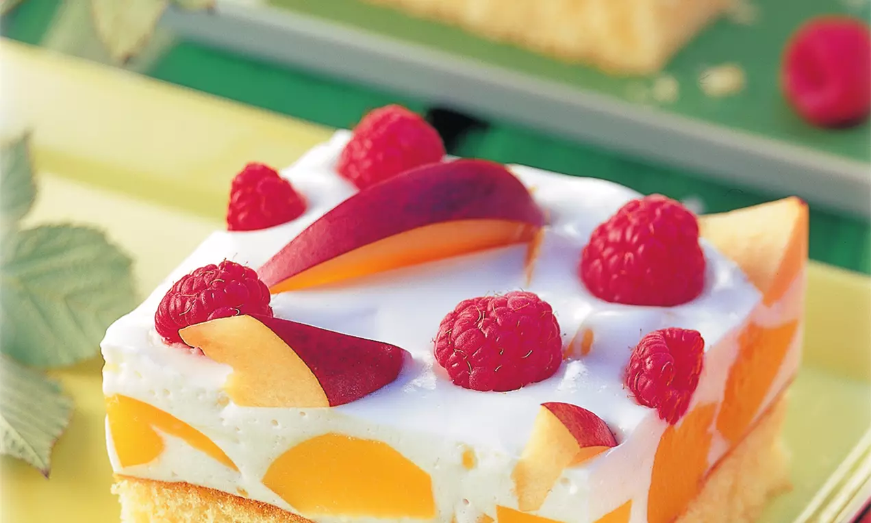 Fruchtkuchen mit hellem Biskuitboden, cremiger weißer Schicht, darin Pfirsichstückchen, garniert mit frischen Himbeeren und Pfirsichspalten