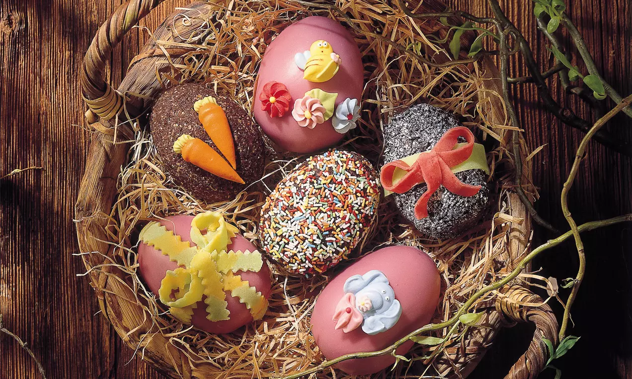 Bunte Ostereier aus Schokolade, verziert mit Zuckerfiguren, Schleifen und Streuseln, gebettet auf strohigem Untergrund in einem Korb