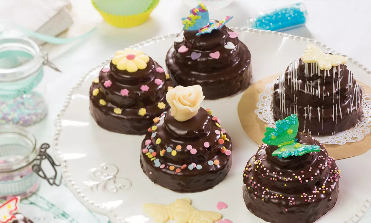 Mini-Marmor-Törtchen mit glänzend dunkler Glasur, dekoriert mit bunten Zuckerstreuseln und filigranen Marzipanfiguren auf einer weißen Platte