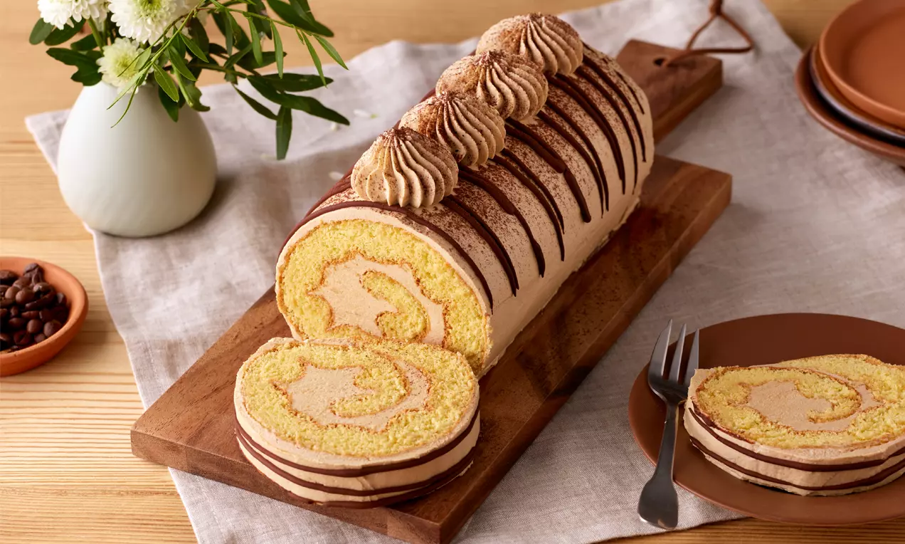 Biskuitroulade mit blassgelber Spirale und sahniger Kaffeecreme, dekoriert mit cremigen Tupfen und Schokostreifen auf Holzbrett angerichtet