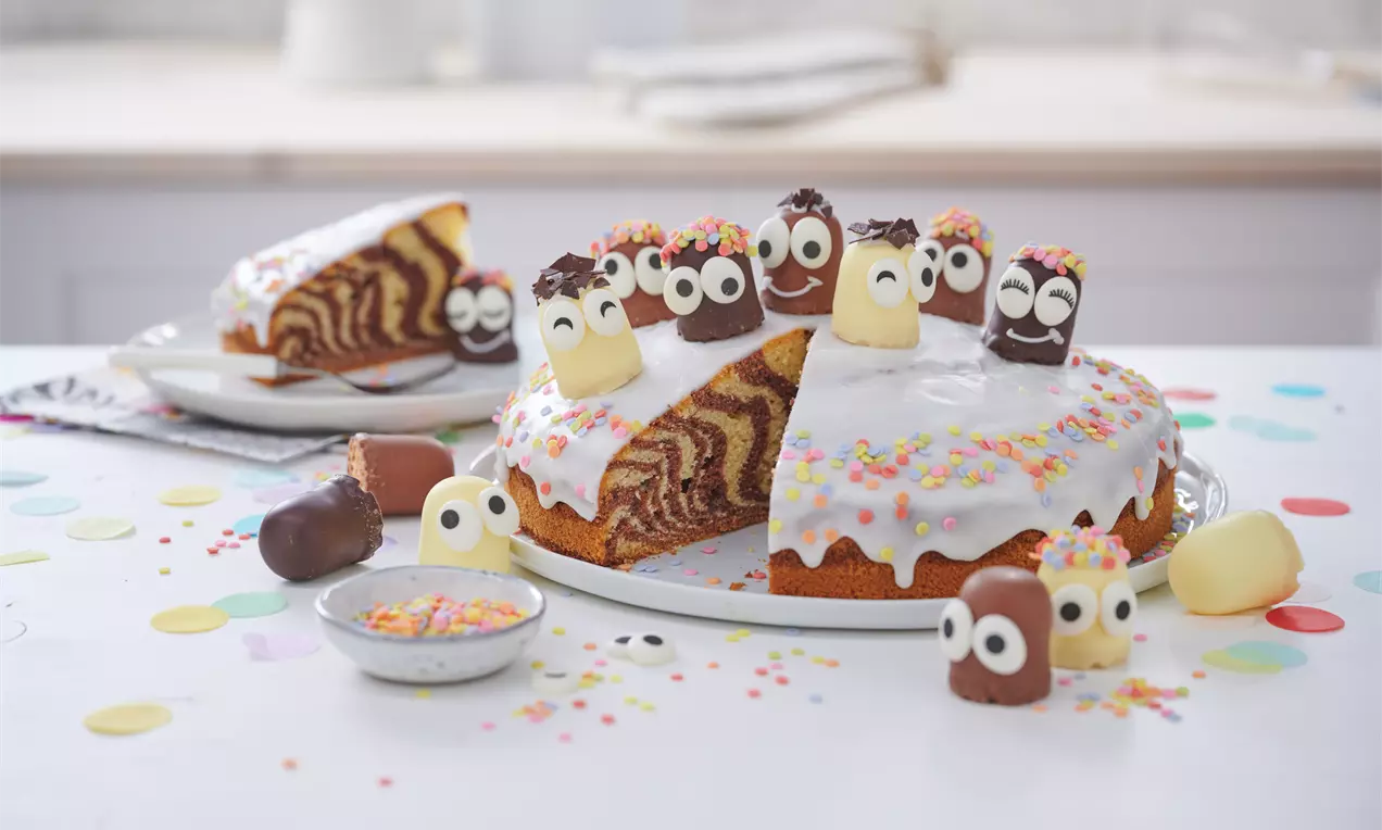 Zebrakuchen mit weißem Zuckerguss, bunten Zuckerstreuseln und verzierenden Schokowürstchen mit Augen auf weißem Teller drapiert