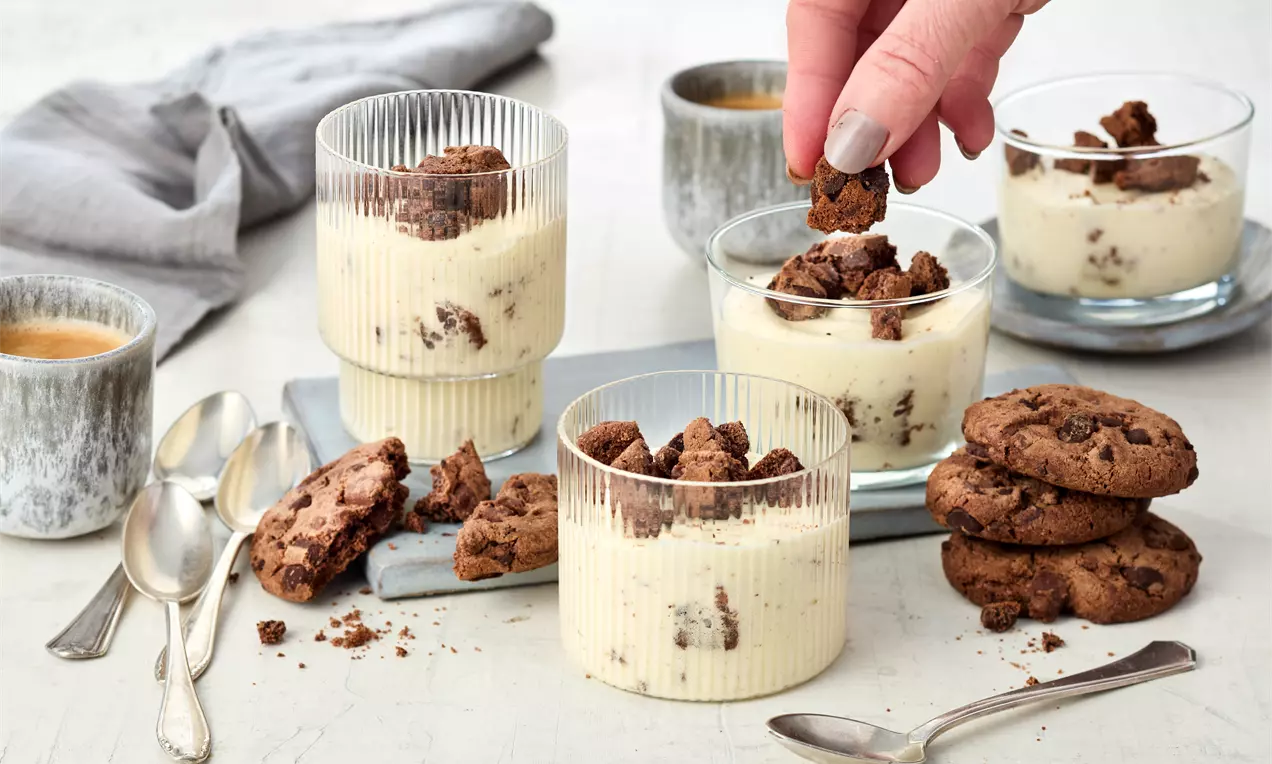 Creme-Dessert in geriffelten Gläsern mit hellbeiger, zarter Textur und knusprigen braunen Cookie-Stücken, angerichtet auf hellem Untergrund