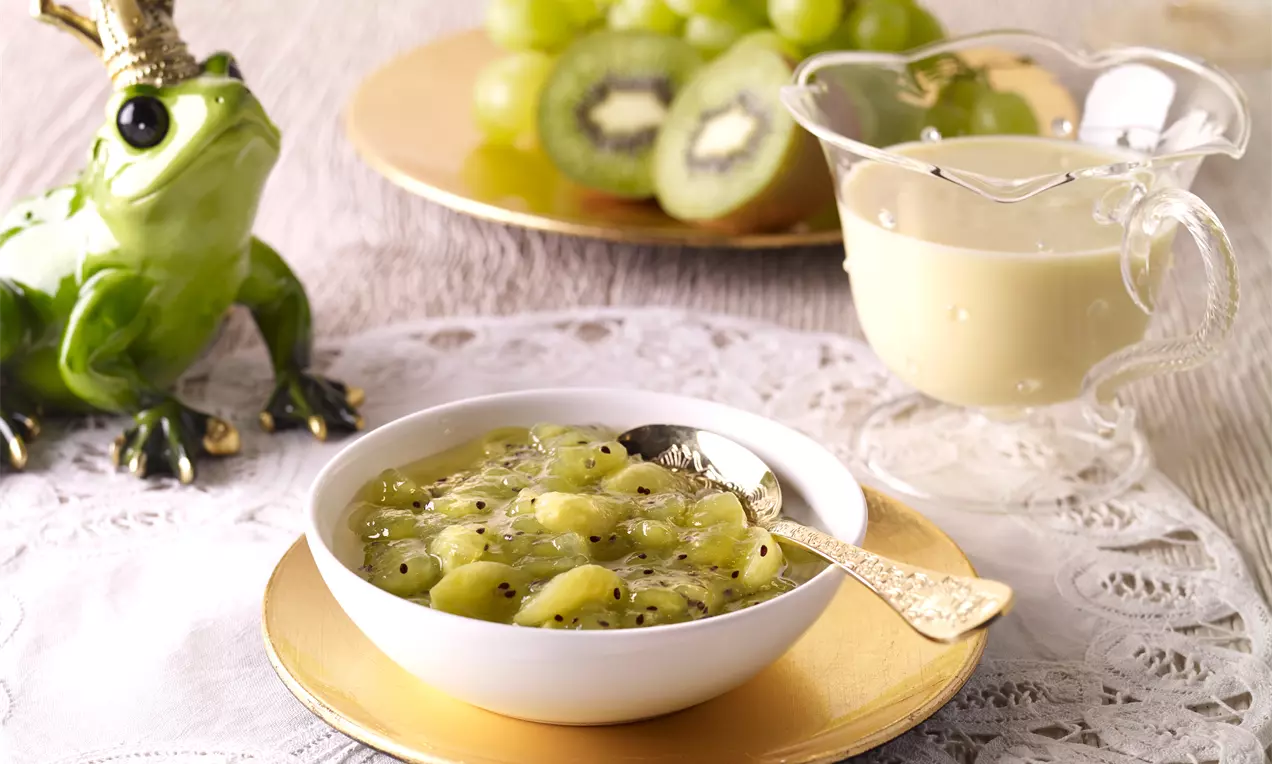 Kiwi-Weintrauben-Grütze mit grüner, glänzender Textur in einer weißen Schale auf einem goldenen Teller mit Sahnekanne und Dekoration
