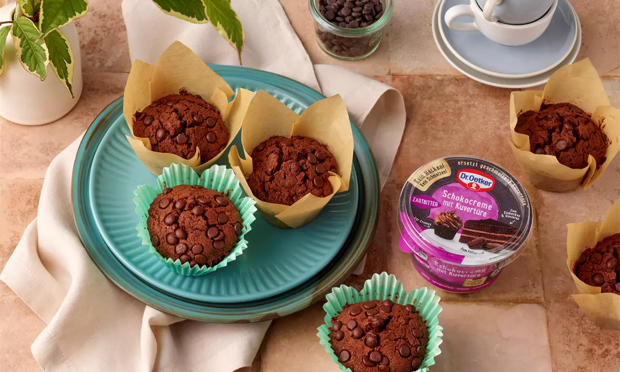 Saftige, dunkelbraune Schokomuffins mit glänzenden Schokodrops in bunten Förmchen und naturfarbenem Papier auf einem türkisfarbenen Teller