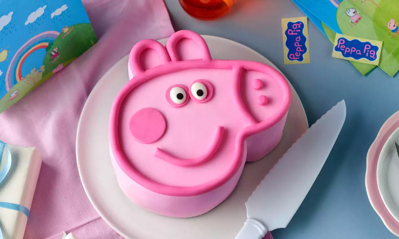 Rosa Peppa-Wutz-Torte in Form eines stilisierten Kopfes mit zarten Konturen, großen Augen und fröhlichem Lächeln auf weißem Teller
