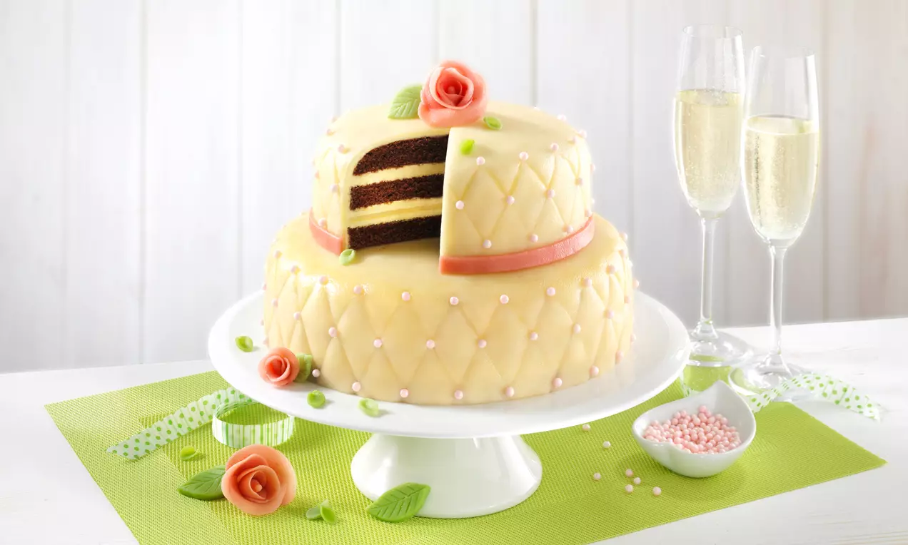 Zweistöckige Marzipantorte mit aprikosenfarbener Oberfläche, zarten Perlen, orangefarbener Borte und marzipanen Rosen auf weißer Tortenplatte