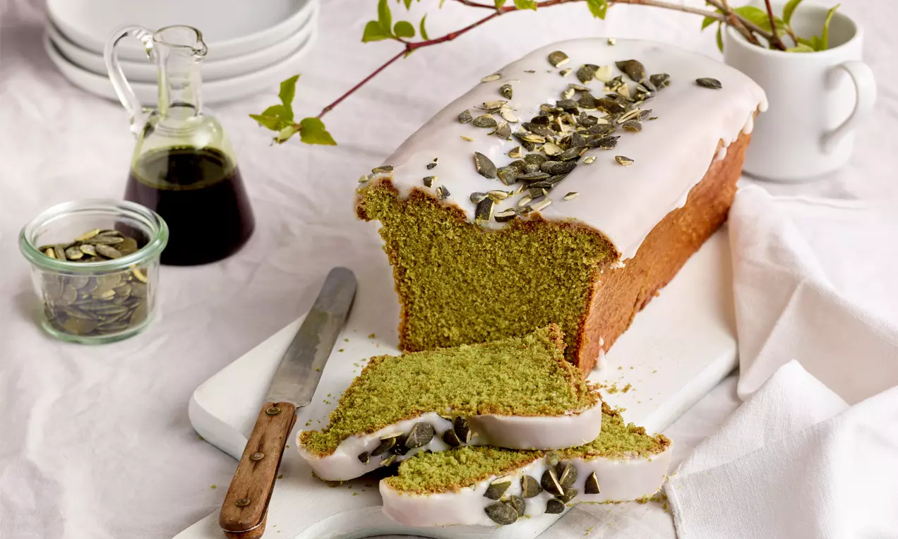 Saftiger Kastenkuchen mit grünem Innenleben, weißem Zuckerguss und Kürbiskernen, angeschnitten auf einem hellen Brett mit Messer drapiert
