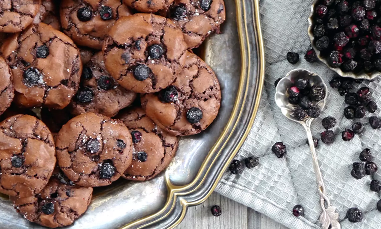 Knusprige, rissige Schoko-Cookies mit dunklen Heidelbeeren, auf einer antiken, silbernen Platte neben getrockneten Heidelbeeren auf Stoff