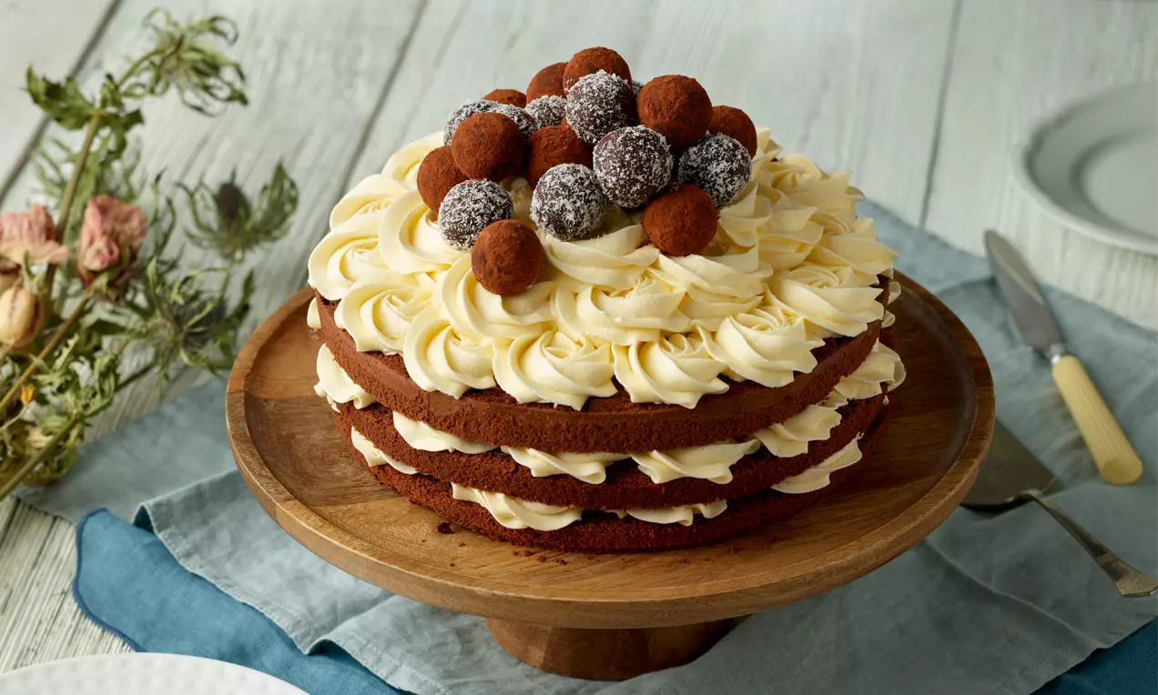 Dreischichtiger Naked Cake mit dunklen Böden, hellgelber Frischkäse-Buttercreme und Kakao-Trüffeln auf einer Holzplatte angerichtet